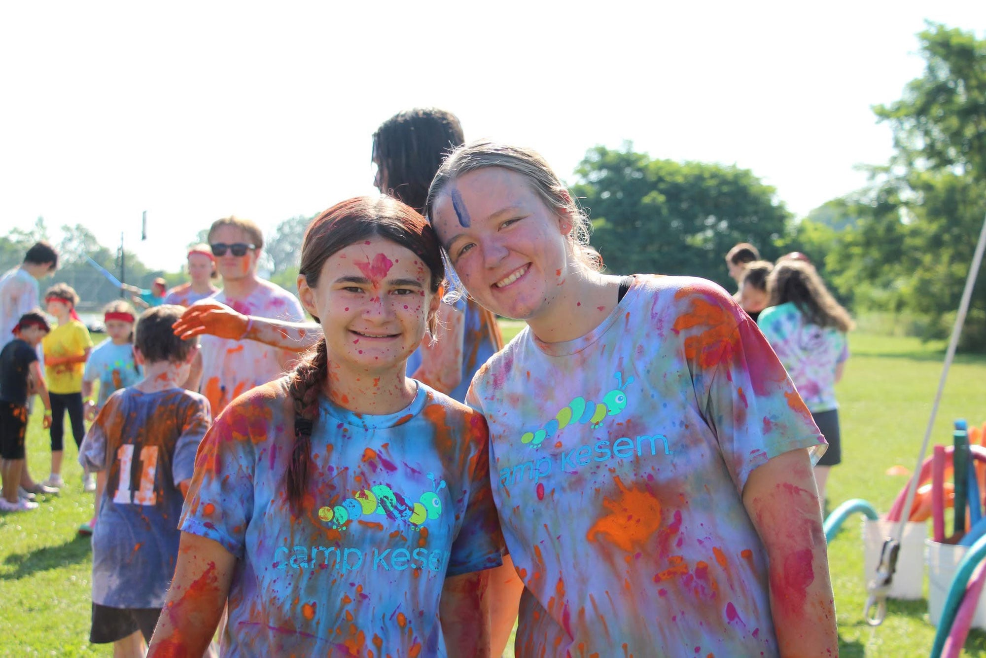 Two young women splattered with paint, standing in a field and smiling at the camera.