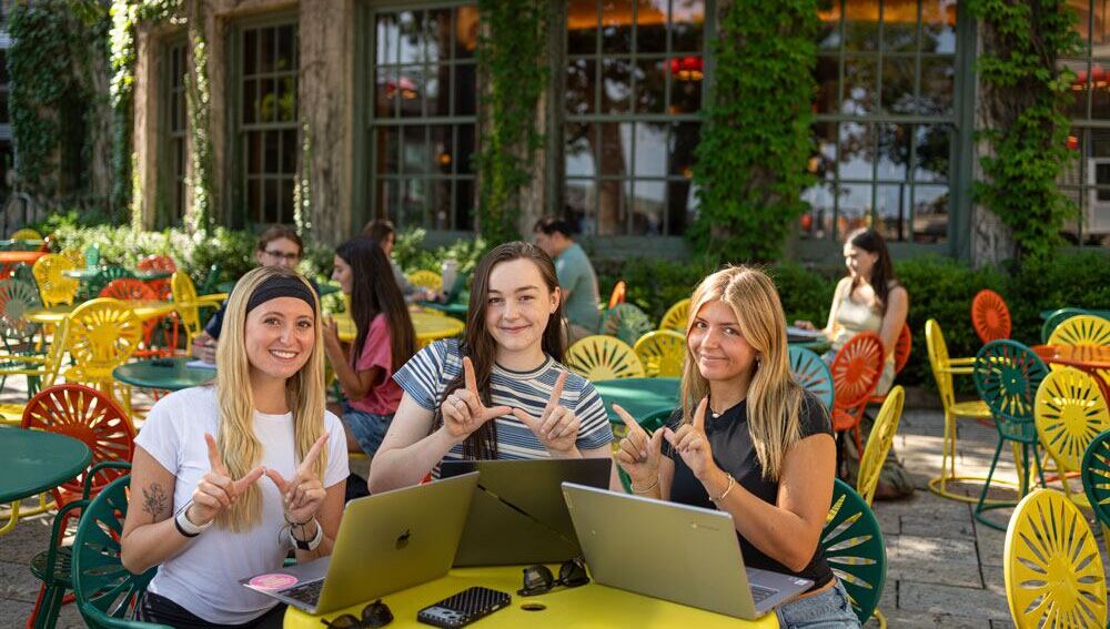 Three students sitting at the Union Terrace, making the UW–Madison 'W' wign
