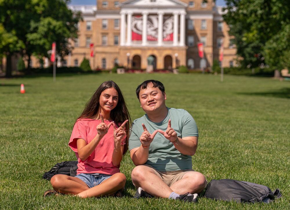 Two summer students making the UW–Madison 'W' hand sign while sitting on the lawn of Bascom Hill