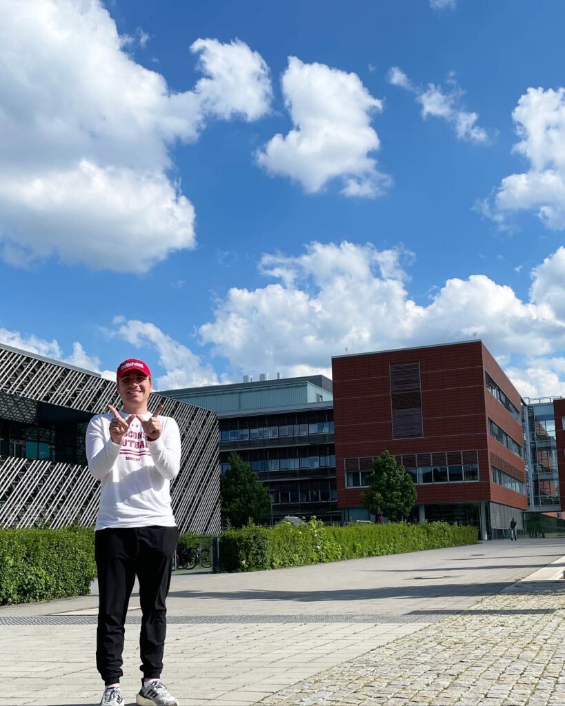 Estudiante usando ambas manos para hacer el signo W frente a un edificio universitario en Alemania