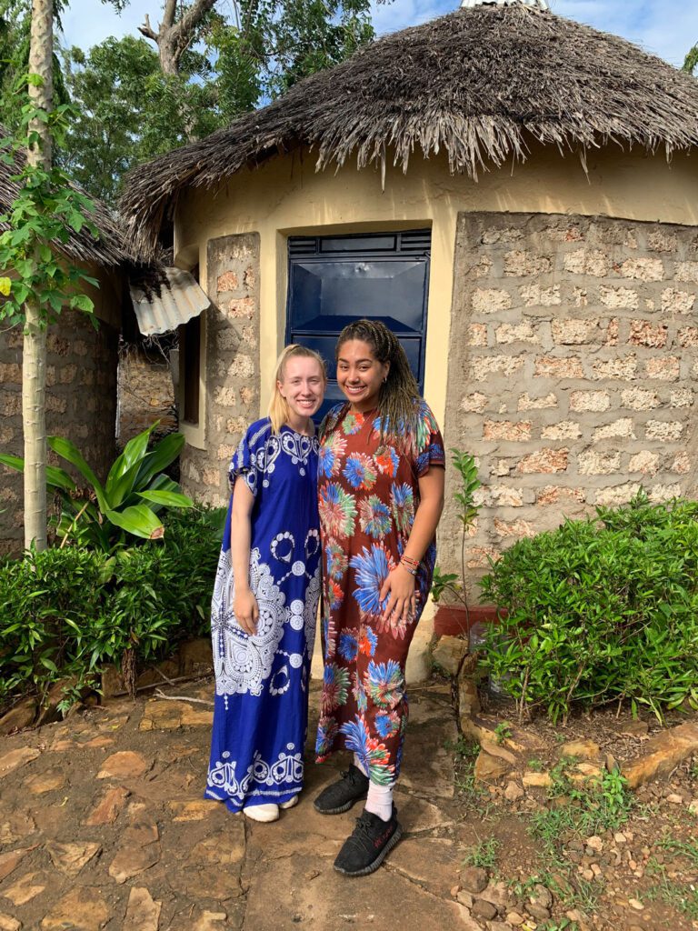 Two students in African prints standing and smiling in front of a small hut