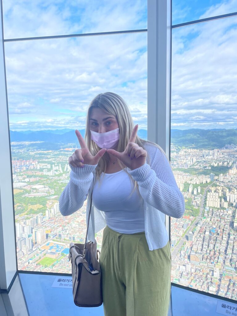 Student standing in front of a window that overlooks Seoul, South Korea, giving the W sign with her hands