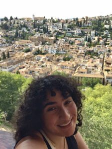Student with curly dark hair smiling with picturesque summer village behind her