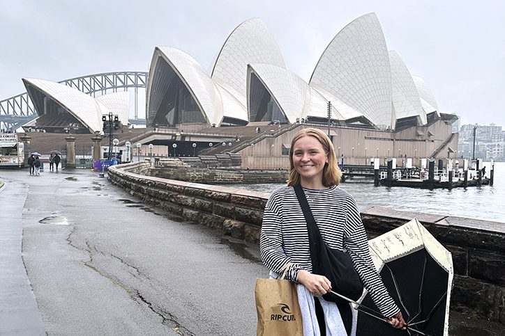 UW–Madison student Maren Stewart stands holding an umbrella to her side on one hand and with a shopping bag in the other. In the background behind her is the Sydney Opera House.