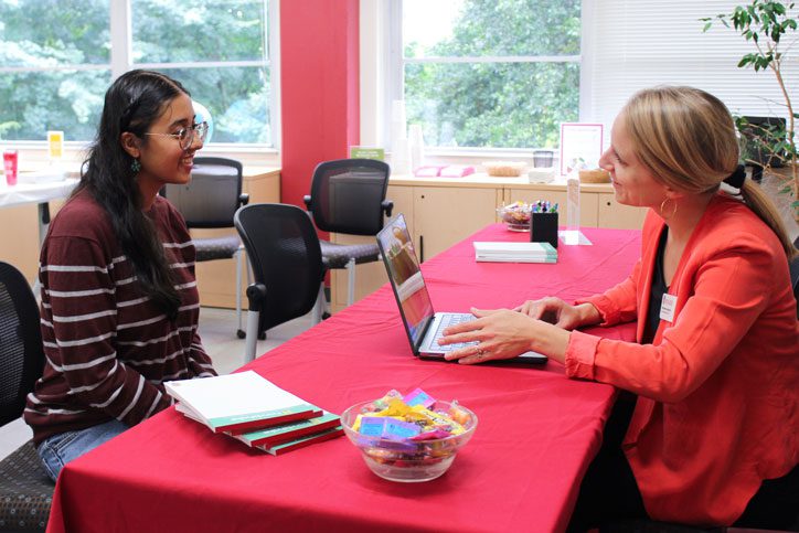 Michelle Jackson sits with her laptop talking to a student across a long table with red tablecloth. Jackson has long blonde hair and is wearing a red blazer. The student has long dark hair and wears glasses and a maroon and white striped sweater.