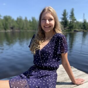 UW–Madison student Sarah Kopp sits with legs outstretched on a lake dock, with water and pine trees blurred behind her. She has long blonde hair and wears a short blue dress with white polka dots.