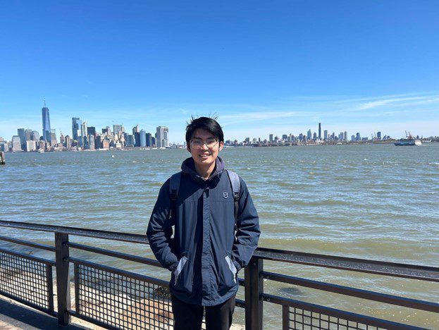 Chentian standing in the new york harbor with the city in his background. he is smiling.