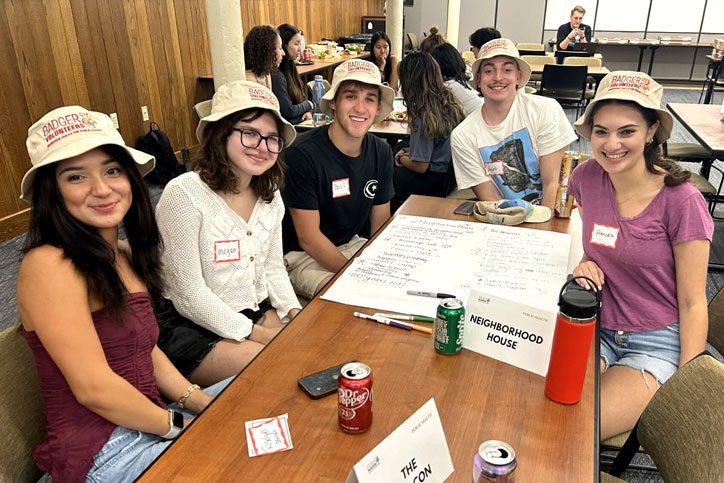Badger Volunteer students sitting around a table, smiling. They are all wearing bucket hats that say "Badger Volunteers"