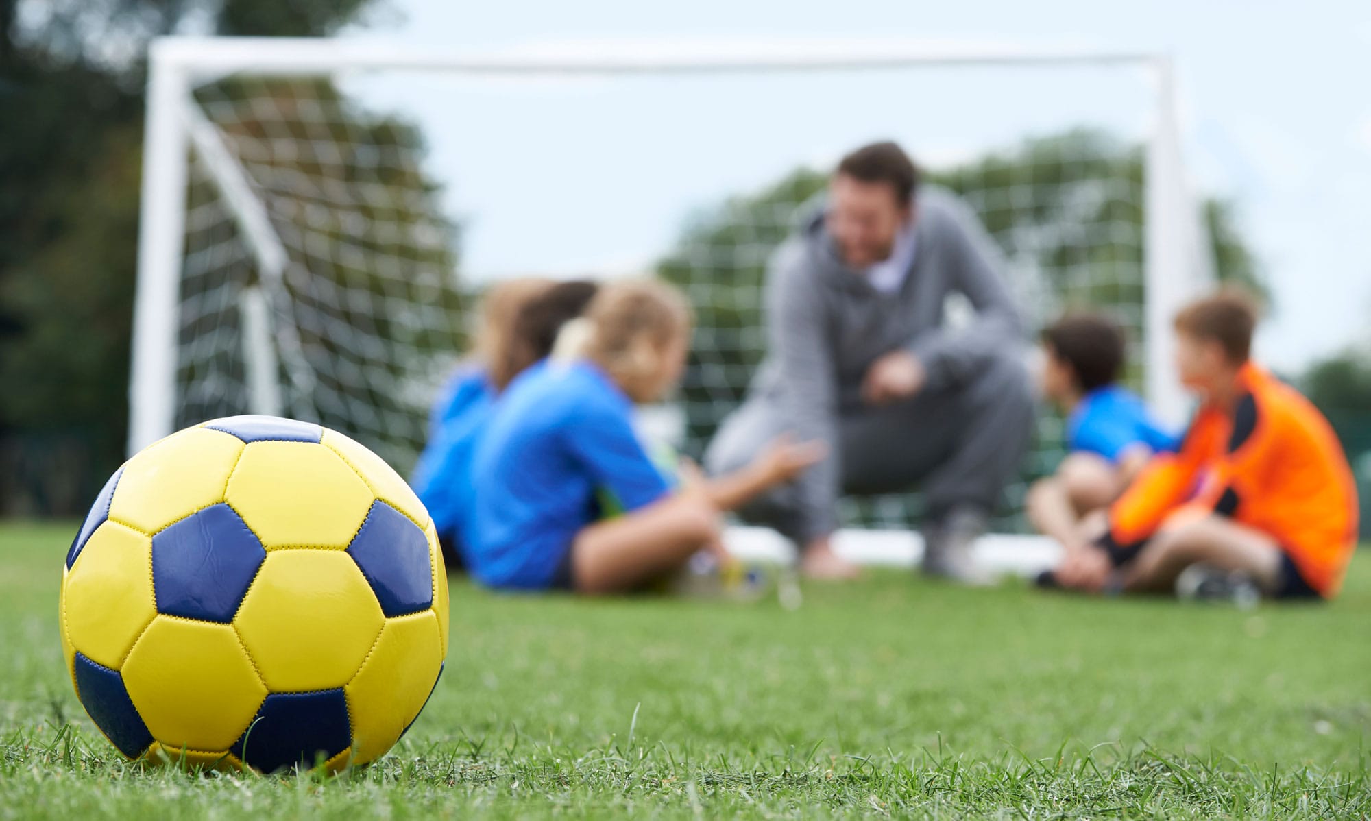 Closeup of a soccer ball in the forefront, while a coach speaks to players in the background