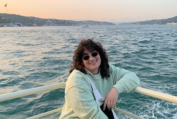 Student sitting at front of boat with bridge and big body if water behind them