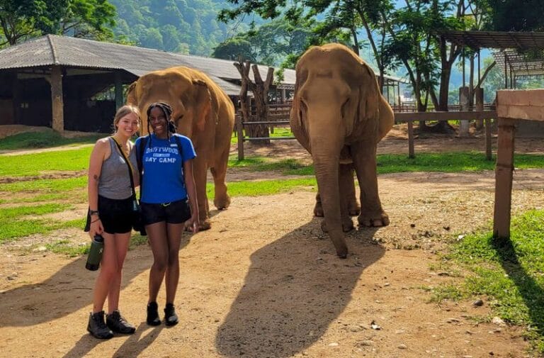 Two students stand facing the camera, smiling for a photo in front of two large elephants. A clear blue sky and large mountain full of lush greenery stand in the background behind students and the elephants.