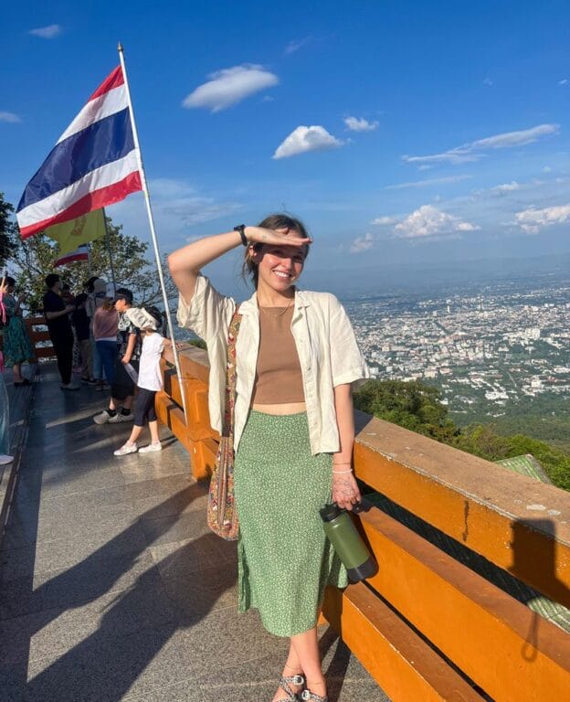 Summer Term student on a hillside in Chiang Mai, Vietnam smiling while blocking the sun with one hand. A Thai flag blows in the breeze in the background.