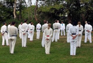 Students wearing white Taekwondo uniforms stand with backs to the camera. They are outdoors, standing on grass with trees in the background, and are listening to instructor, shown in the background.