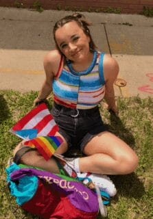 Isa Hernandez-White sits in the grass in front of a sidewalk, dressed in bright colors with pride flag and Puerto Rico flag in her lap