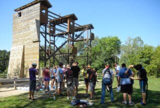 A group of high school and college students prepare to go on a low ropes course. A low ropes tower is seen in the background, with wood surrounding the area.