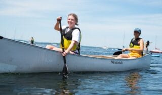 Two students in a canoe on the water smile at the camera. Both are wearing life jackets and paddling. Sailboats and trees are in the background.