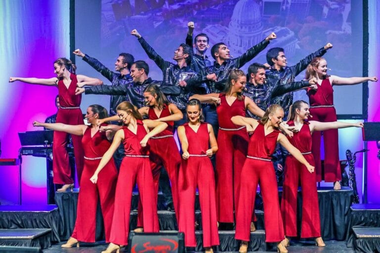 UW–Madison Wisconsin Singers stand smiling in bright costumes with arms outstretched in various directions to form a starburst pattern.