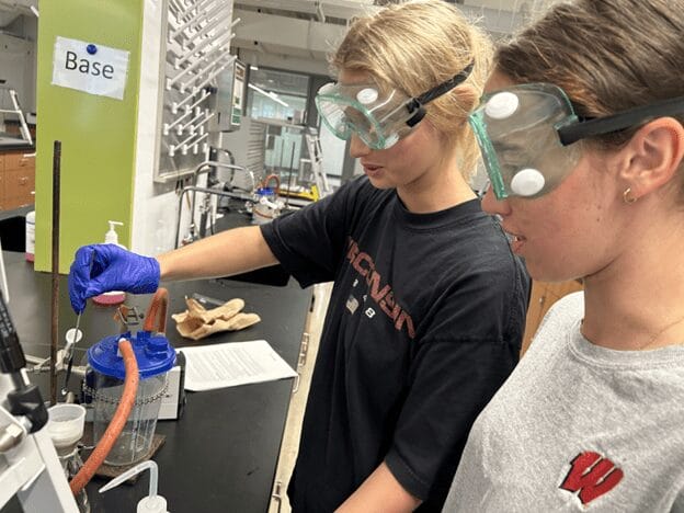 Two women students in a chemistry lab wearing goggles and working with lab equipment