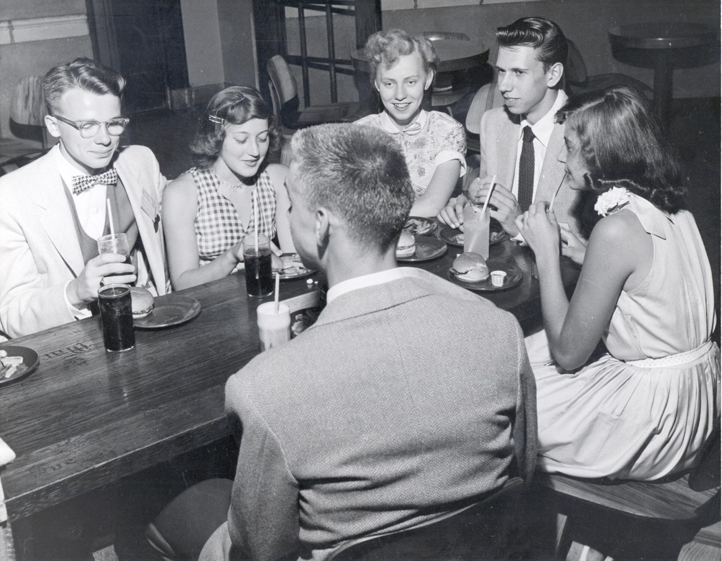 Six young men and women in suits and dresses sit around a table in the Rathskeller, drinking sodas and eating burgers.