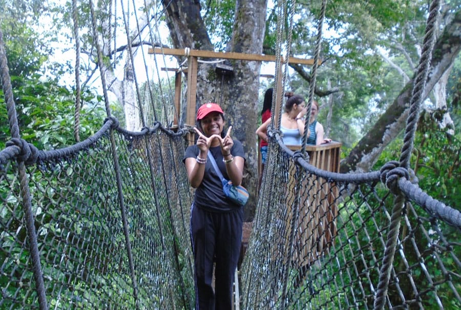 Naureen Kamal stands on a rope bridge in the tree tops while making the “W” sign with her fingers.